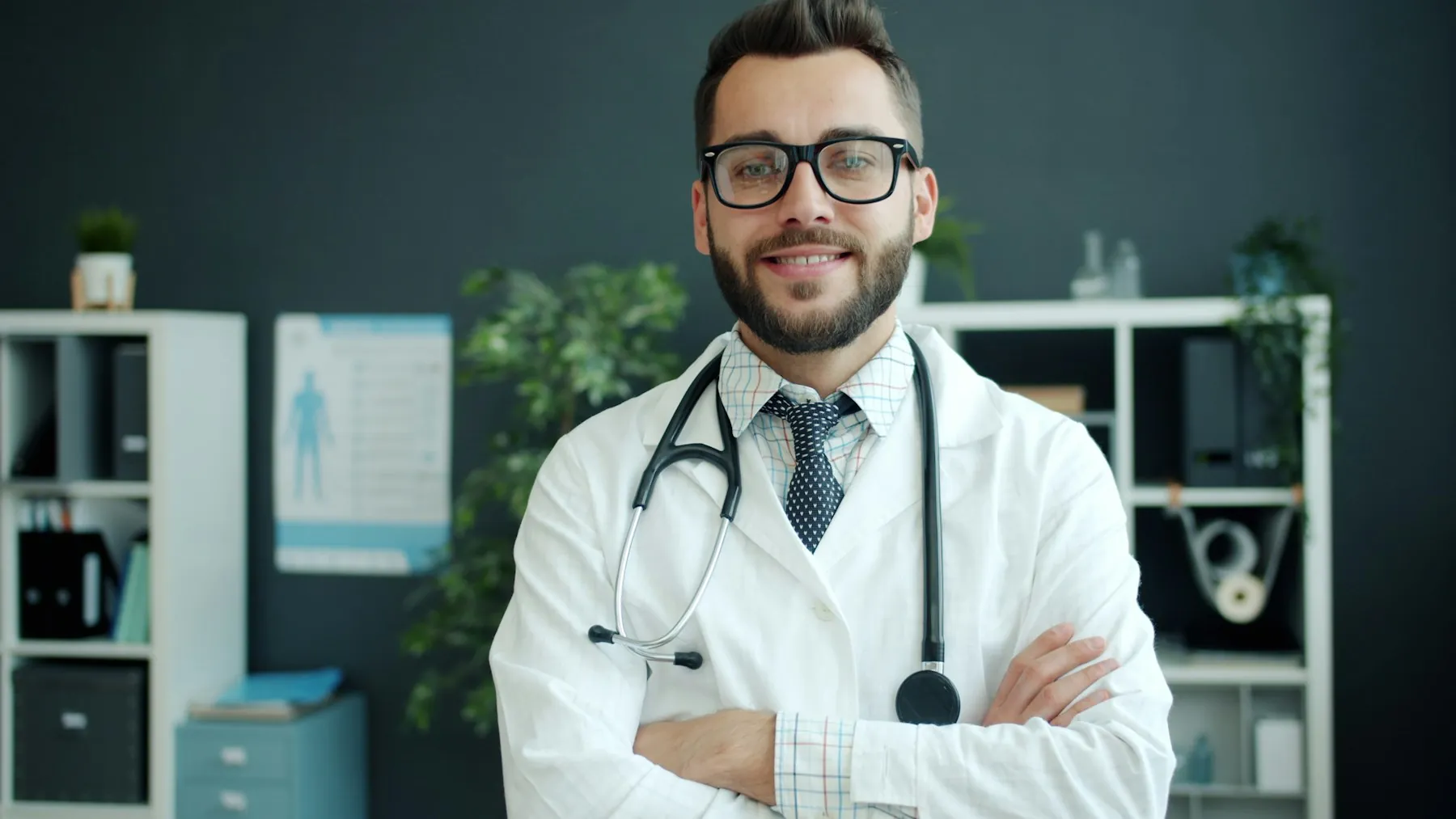 A smiling doctor with glasses and a stethoscope standing with arms crossed in a consulting room